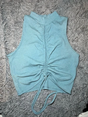 Blå ribbad croppad linnetopp från Timing - Säljer en blå croppad linnetopp från Timing med hög hals och snygg snörning framtill. Toppen är ribbad och ärmlös, vilket ger en cool och modern vibe. Perfekt för varma dagar och att styla med jeans eller kjol.