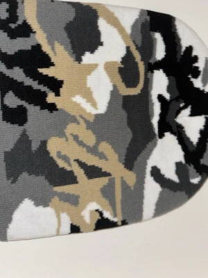 Cortez mössa - Säljer en stickad väst med coolt camouflage-mönster i svart, grått, beige och vitt. Västen har en klassisk rak form och är perfekt för dig som vill sticka ut med en streetstyle-look. Materialet är mjukt och bekvämt stickat tyg.