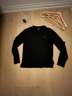 Svart långärmad tröja från Polo Ralph Lauren - Svart långärmad tröja från Polo Ralph Lauren med klassisk röd logobrodering på bröstet. Tröjan har rund hals och är gjord i mjuk bomull, perfekt för en chill och stilren look. Passformen är normal och materialet känns skönt mot huden.