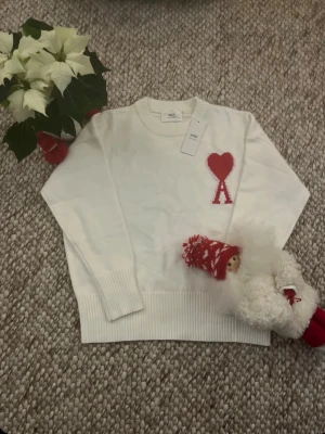 Vit Ami Paris stickad tröja - Säljer en vit Ami Paris stickad tröja med röd hjärtapplikation och bokstaven A på bröstet. Tröjan har ribbade muddar, rund hals och en clean, oversized passform. Perfekt statement-piece för vintern och julen. St-S fast passar även m. Skick 10/10 priset går att ändras.