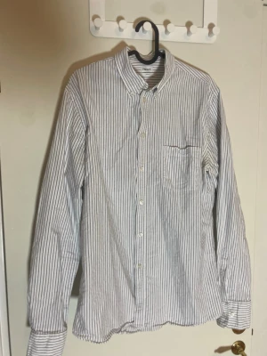 Randig skjorta från Filippa K, M - Klassisk vit skjorta med smala svarta ränder från Filippa K. Skjortan har lång ärm, bröstficka och knäppning framtill. Perfekt för dig so gillar stilren och enkel design. Tillverkad i bomull för en skön känsla.