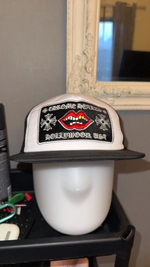 Chrome Hearts truckerkeps svart/vit - Säljer en fet truckerkeps från Chrome Hearts i svart och vitt med mesh på sidorna och bak. Framsidan har ett broderat motiv med röda läppar och tänder, samt texten 'Hollywood USA'. Kepsen har justerbar snapback och coola detaljer som metallknapp och broderade band invändigt.
