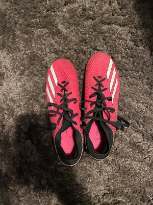 Adidas rosa fotbollsskor med dobbar - Säljer ett par snygga fotbollsskor från Adidas i stark rosa färg med svarta och vita detaljer. Skorna har klassiska tre ränder på sidan, snörning och en ovandel i syntetmaterial. Yttersulan har svarta dobbar för bra grepp på planen. Perfekt för dig som vill sticka ut på matchen!