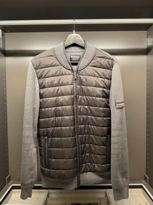 Barbour cardigan - Grå cardigan från Barbour med quiltad front och gråa ärmar. Jackan har ribbade muddar, dragkedja framtill och två fickor med zip.