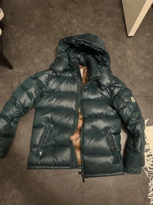 Moncler Maya - Säljer en mörkgrön pufferjacka från Moncler med huva och bruna detaljer på insidan. Jackan har två dragkedjeförsedda fickor, quiltad design och Moncler-logga på ärmen. Perfekt för kalla dagar och har en snygg, modern look. Storlek 2. Passar S/M