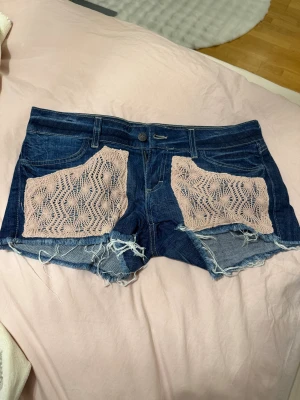 Lågmidjade shorts - Snygga blå jeansshorts med ljusrosa spets på framfickorna och råa, fransiga kanter. Baksidan har två fickor med broderade mönster. Perfekta för dig som gillar en chill och unik stil med extra detaljer. Pris går att diskutera💕