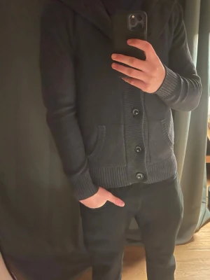 Mörkgrå stickad fodrad Cardigan  - Säljer en mörkgrå fodrad cardigan i storlek medium. Cardiganen har huva, långa ärmar och knäppning med fyra knappar framtill. Den är stickad och har två fickor på sidorna samt ribbade muddar vid ärmslut och nederkant. Perfekt för kyliga dagar.