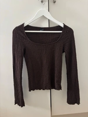 Brun ribbad topp från Gina Tricot - Mörkbrun långärmad topp från Gina Tricot i ribbat, stretchigt material. Toppen har en rund, djup ringning och smal passform som sitter snyggt på kroppen. Perfekt att styla med jeans eller kjol för en enkel men trendig look.