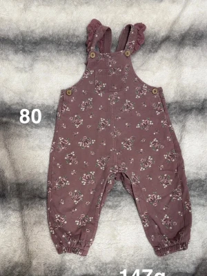 Blommig hängselbyxor med volangaxel - Supersöta hängselbyxor med blommigt mönster och volangdetaljer på axelbanden. Hängselbyxorna har knappar vid axlarna och en ficka framtill. Resår vid bensluten för skön passform. Perfekt för dig som gillar gulliga och lekfulla plagg!🌸 Fint använt skick. Storlek 80