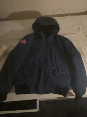 Mörkblå Canada Goose dunjacka - Säljer en mörkblå dunjacka från Canada Goose med huva och avtagbar ljus pälsdetalj. Jackan har klassisk patch på ärmen och är riktigt varm med quiltad insida. Perfekt för kalla vinterdagar och har en stilren look.