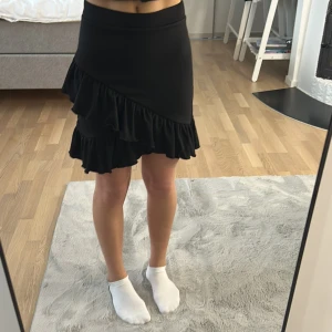 Svart volangkjol från Nelly XS - Supersöt svart kjol från Nelly med volangdetaljer framtill. Kjolen har struktur i tyget och resår i midjan. Aldrig använd men klippt bort lappar. 