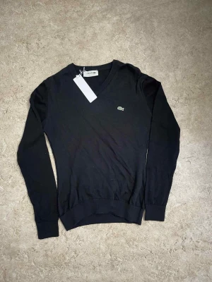 Svart v-ringad tröja från Lacoste - Klassisk svart v-ringad tröja från Lacoste med den ikoniska gröna krokodilloggan på bröstet. Tröjan har lång ärm och ribbade muddar vid ärmslut och nederkant. Perfekt för dig som gillar stilrena och tidlösa plagg.