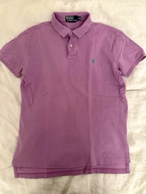 Lila pikétröja från Polo Ralph Lauren - Säljer en lila pikétröja från Polo Ralph Lauren i storlek M. Tröjan har klassisk krage, korta ärmar och två knappar vid halsen. Ikoniska gröna loggan broderad på bröstet. Perfekt för dig som gillar stilrena och färgglada plagg.
