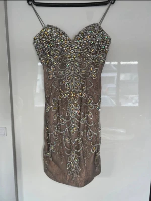 Kort glittrig klänning med paljetter/stenar - Supersnygg kort klänning med tunna axelband i beige/brun ton, täckt av glittriga paljetter och stenar i ett blommigt mönster. Klänningen har en sweetheart-ringning och är perfekt för dig som vill sticka ut på festen. Materialet känns lyxigt och skimrande.