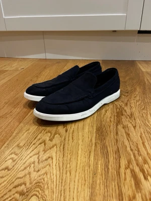 Mörkblå loafers med vit sula - Snygga mörkblå loafers med klassisk design och vit platt sula. Skorna är tillverkade i mocka och har en stilren, enkel look utan snörning. Perfekta för dig som gillar en clean och avslappnad stil. Har använt dom 2 gånger 