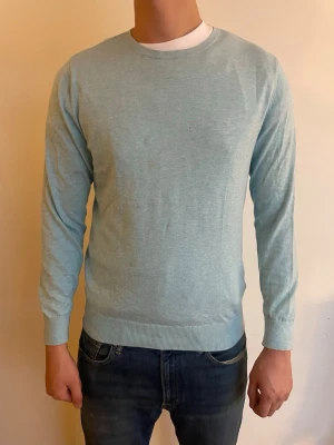 Ljusblå långärmad tröja - Säljer en ljusblå tunn tröja med rund hals och långa ärmar. Tröjan har ribbade muddar vid ärmslut och nederkant, perfekt för lager på lager under vår och höst. Enkel och stilren design som passar till jeans eller chinos.