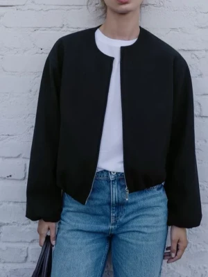 Zara bomberjacka - Säljer en svart bomberjacka med enkel, stilren design och rundad halsringning. Jackan är utan synliga knappar eller dragkedja framtill och har långärmade ärmar. Perfekt för dig som gillar en clean och modern look. Från Zara. Helt ny!!  Passar S och M