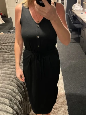 Svart ärmlös klänning med knappar - Svart klänning med v-ringning och knappar framtill. Ärmlös modell med knyt i midjan och slits på sidan. Klänningen har en rak, enkel siluett och är i midi-längd. Perfekt för dig som gillar stilrena och bekväma plagg.