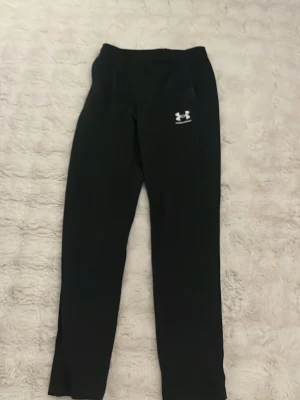Svarta mjukisbyxor från Under Armour - Svarta mjukisbyxor från Under Armour med logga på vänster lår. Byxorna har elastisk midja och raka ben, perfekta för chill eller träning. Tillverkade i mjukt och bekvämt material som andas. Klassisk och enkel design utan extra detaljer.