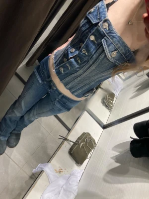 Jeans-korsett med knappar - Säljer en  jeanskorsett i blå denim med silverfärgade knappar framtill och dekorativa fickor. Den har blivit lite glittrig men inget som man märker av. 