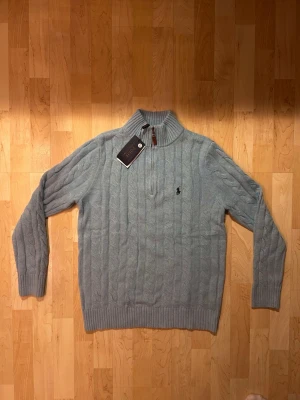 Ljusblå stickad tröja Polo Ralph Lauren - Säljer en ljusblå stickad tröja från Polo Ralph Lauren med kabelstickat mönster och hög krage. Tröjan har half zip-dragkedja med läderdetalj och broderad logga på bröstet. Perfekt för kyliga dagar och riktigt snygg till jeans.