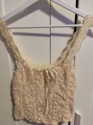 Beige spetsig linnetopp med rosett - Säljer en superfin beige linnetopp med spetsdetaljer och breda axelband. Toppen har ett vackert blommigt spetsmönster, dekorativ rosett framtill och tunna band längs sömmarna. Står inte storlek men skulle säga M