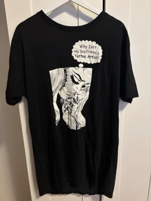 Svart t-shirt med print - Svart t-shirt från Koi Tattoo med stort grafiskt tryck fram på en tecknad tjej och texten 'Why isn't my boyfriend a Tattoo Artist?'. Baksidan har en stor logga i vit, snirklig stil. Klassisk passform och rund hals.