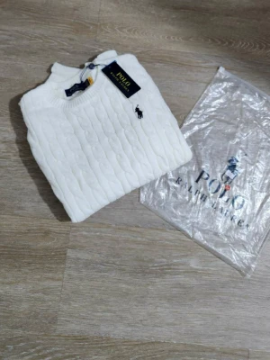 Vit kabelstickad tröja Polo Ralph Lauren - Vit kabelstickad tröja från Polo Ralph Lauren med klassisk rund halsringning och svart broderad logga på bröstet. Tröjan har ribbade muddar och är långärmad, perfekt för en clean och stilren look.