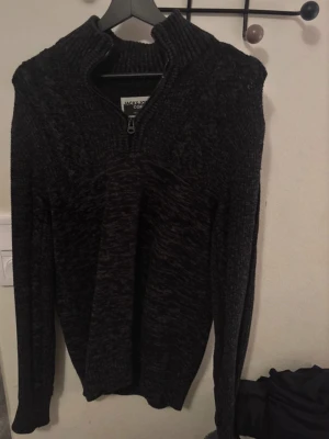 Mörk stickad tröja från Jack & Jones - Säljer en mörkgrå stickad tröja från Jack & Jones Core med half zip-dragkedja i halsen. Tröjan har lång ärm och en hög krage som ger extra värme. Perfekt för kyliga dagar och enkel att matcha med jeans eller chinos.