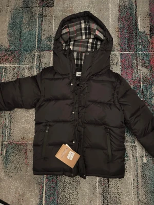 Svart pufferjacka från Burberry S - Säljer en svart pufferjacka från Burberry med huva och rutigt foder i svart, vitt och rött. Jackan har dragkedja, knappar och två fickor framtill. Klassisk Burberry-logga på ärmen. Perfekt för kalla dagar och riktigt snygg streetstil.