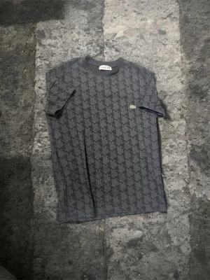 ÄKTA mönstrad  t-shirt från Lacoste - Säljer en svart t-shirt från Lacoste i storlek S med geometriskt mönster över hela tröjan och klassisk krokodil-logga på bröstet. Tillverkad i mjuk bomull med lite stretch för extra komfort. Perfekt för dig som gillar stilrena men ändå unika plagg.
