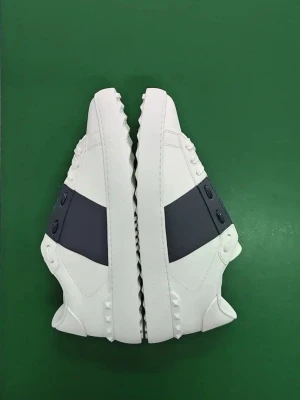 Valentino Garavani vita sneakers - Säljer ett par snygga vita Valentino Garavani sneakers med bred mörkblå rem över ovandelen och coola nitar runt hälen. Skorna är i skinn och har klassisk rund tå och platt sula. Perfekta för dig som vill ha en stilren men edgy look.