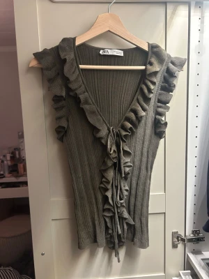 Olivgrön ribbstickad med vollang detaljer och knytning fram till - ZARA ribbstickad topp i olivgrön nyans. Perfekt till en utekväll eller en fin middag. Volanger framtill och vid ärmar ger en elegant look. Med knytning framtill. Storlek M.