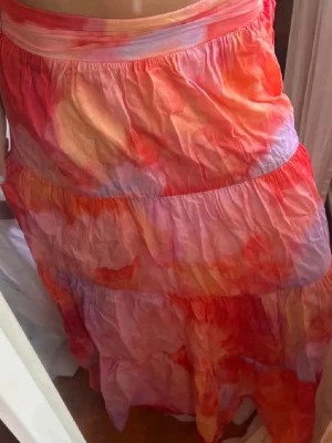 Färgglad långkjol från Lindex  - Säljer en flowy långkjol i mjuk bomull med färgstarkt mönster i rött, orange, rosa och lite lila. Kjolen har flera volanger och en bred midjeresår som gör den superbekväm. Perfekt för dig som gillar boho-vibbar och vill sticka ut lite extra.