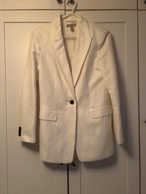 Vit kavaj i linne från H&M - Snygg vit kavaj från H&M i linne med klassisk krage och en knapp framtill. Två fickor med lock och svarta knappar vid ärmsluten. Perfekt för dig som vill ha en stilren och fräsch look.