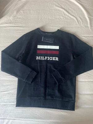 Svart sweatshirt från Tommy Hilfiger - Svart sweatshirt från Tommy Hilfiger med klassisk logga och färgblock i vitt, rött och marinblått på bröstet. Tröjan har rund hals, långa ärmar och ribbade muddar. Perfekt för dig som gillar stilrena och ikoniska märken. Pris kan diskuteras vid seriösa bud
