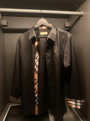 Svart Burberry skjorta med rutiga detaljer - Säljer en svart skjorta från Burberry med klassiska beige-rutiga detaljer längs knappslån och insidan av manschetterna. Skjortan har lång ärm, krage och knappar framtill. Materialet känns mjukt och exklusivt, perfekt för dig som vill ha något stilrent men ändå med en twist. (SITTER Slim)