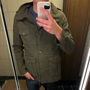 Olivgrön parkajacka från H&M - Säljer en olivgrön parkajacka från H&M med fyra stora fickor framtill, axelklaffar och knappar. Jackan har dragsko i midjan och huva, perfekt för dig som vill ha en stilren och funktionell jacka till vardags. Tillverkad i bomullsmix.