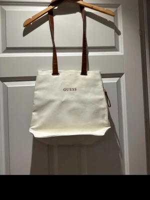 Vit ryggsäck från Guess med bruna detaljer - Snygg och stilren ryggsäck från Guess i vit bomull med bruna axelband i skinnimitation. Väskan har dragkedja på sidan och enkel design med Guess-logga framtill. Perfekt storlek för vardagens alla grejer och lätt att matcha till olika outfits.