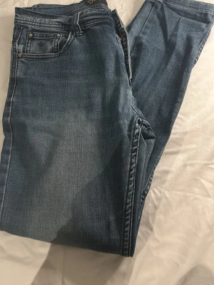 Blå jeans från Jokob JNS - Säljer ett par klassiska blå jeans från Jokob JNS med fem fickor och snygga slitningar. Jeansen har raka ben och silverfärgade nitar samt knapp med logga. Perfekta till sneakers eller boots och har en tidlös tvättad look.