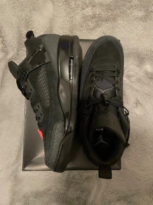 Svarta Air Jordan 4 Spizike sneakers - Säljer ett par svarta Nike Air Jordan 4 sneakers med snygga detaljer i mocka och skinn. Skorna har klassisk snörning, meshpaneler på sidorna och coola loggor på hälen. Perfekta för dig som vill ha en stilren och ikonisk look.
