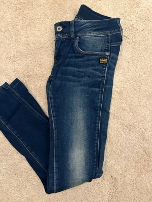G-Star Raw blå lågmidjade jeans - Säljer ett par blå lågmidjade jeans från G-Star Raw med snygga slitningar och klassisk femficksdesign. Jeansen har en liten dragkedja på bakfickan