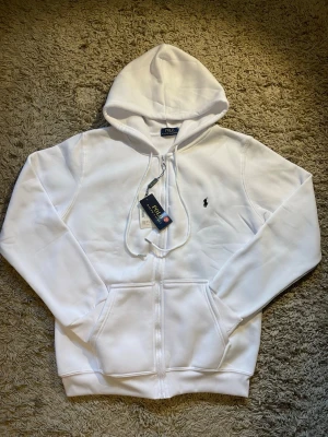 vit hoodie från Polo Ralph Lauren - Säljer en stilren vit hoodie från Polo Ralph Lauren med dragkedja och klassisk huva. Tröjan har det ikoniska broderade logotypen på bröstet och praktiska fickor framtill. Perfekt för dig som gillar en clean och enkel look.