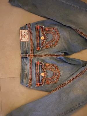 Ovanliga True Religion jeans - Säljer ett par blå True Religion jeans med ikoniska fickor och orange kontrastsömmar. Klassisk femficksmodell med snygga detaljer och logga bak. Jeansen har bootcut-snitt och är tillverkade i jeansmaterial med coola knappar och broderade ficklock. W26. Nypris 2300kr. De har slitningar längst ned men inget man märker av när man bär dem.