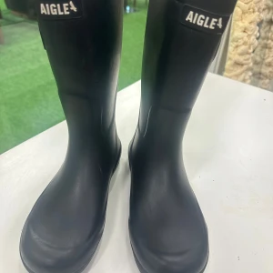 Svarta gummistövlar från Aigle - Svarta höga gummistövlar från Aigle med justerbart spänne på sidan. Klassisk design med rund tå och grov sula för bra grepp. Insidan är ljus och fodrad för extra komfort. Perfekta för regniga dagar och utomhusaktiviteter.