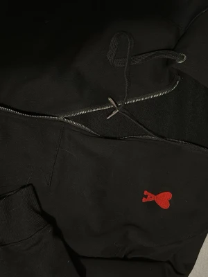 Svart hoodie från Ami med röd logga - Säljer en svart hoodie från Ami Alexandre Mattiussi med dragkedja och huva. Tröjan har en ikonisk röd hjärta-A brodyr på bröstet och ribbade muddar. Perfekt för dig som gillar stilrena detaljer och streetwear-vibe Använd 2-3 gånger i utmärkt skick utan hål eller smuts. priset kan diskuteras!