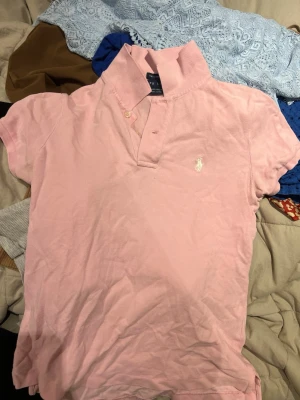 Rosa pikétröja från Ralph Lauren - Säljer en klassisk rosa pikétröja från Ralph Lauren med korta ärmar och vit broderad logga på bröstet. Tröjan har krage, knappar vid halsen och är gjord i mjuk bomull. Perfekt till jeans eller kjol för en sportig look. Vet inte om den är äkta då jag köpt den second hand !!!