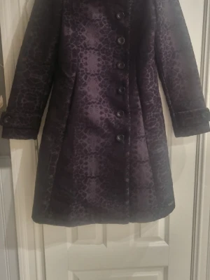 Svart kappa från Desigual - Snygg Svart kappa från Desigual med leopardmönster och stora knappar framtill. Jackan har en klassisk kappa-form med lång ärm och dekorativa detaljer vid ärmslut. Materialet känns mjukt och lyxigt, perfekt för dig som vill sticka ut med en unik look.