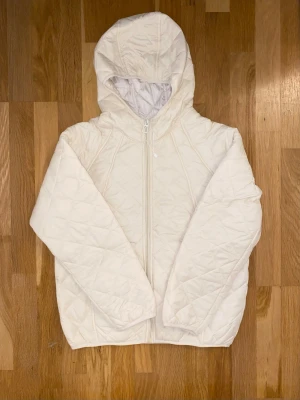 Beige quiltad pufferjacka med huva - Säljer en beige quiltad pufferjacka med huva och dragkedja framtill. Jackan har ett stilrent rutmönster och är tillverkad i lätt polyester, perfekt för kyliga dagar. Långärmad modell med normal passform och enkel, clean look.