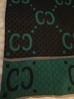 Svart och grön halsduk med CC-mönster - Säljer en unik halsduk i svart och grön färg med stort CC-mönster och små loggor över hela tyget. Västen har fransad kant nedtill och är tillverkad i ett mjukt, syntetiskt material. Perfekt för dig som vill sticka ut med ett coolt och grafiskt mönster.